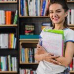 Jovem estudante segurando cadernos em frente estante de livros