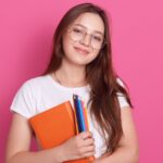 Menina estudante com óculos, caderno e lápis em fundo rosa