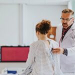 Mulher de costas em roupa hospitalar e homem a examinando com estetoscópio