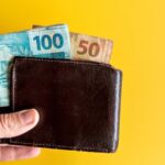 Pessoa segurando carteira com notas de cem e cinquenta em fundo amarelo